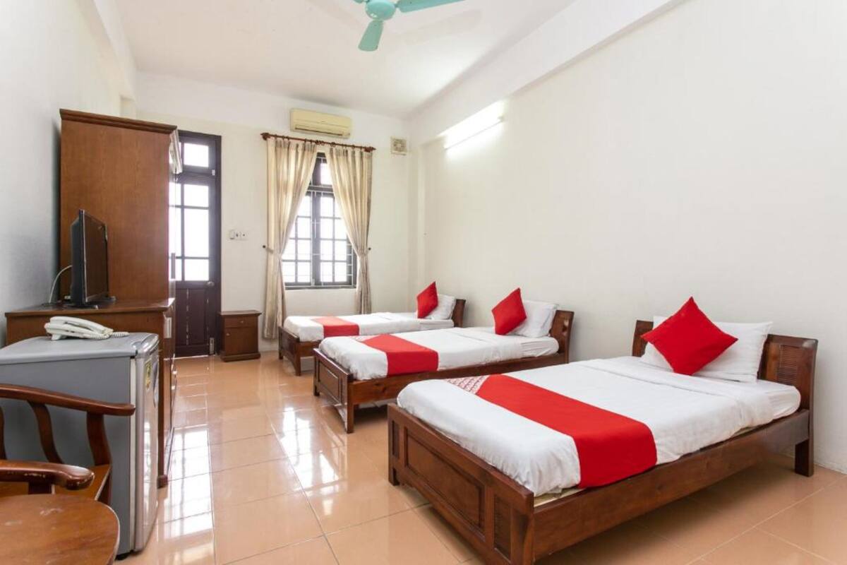 A hotel room with three wooden beds, each with white and red bedding, a desk with a telephone, and a ceiling fan.