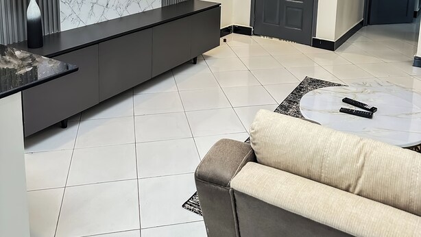 A modern living room with a flat-screen TV mounted on the wall, a dark grey sofa, a marble-patterned coffee table, and a tiled floor. - LivLuxe Apartments 3 (Kampala)