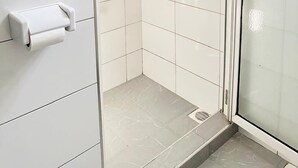 A shower with a glass door, white tiled walls, and a toilet paper holder. - LivLuxe Apartments 3 (Kampala)