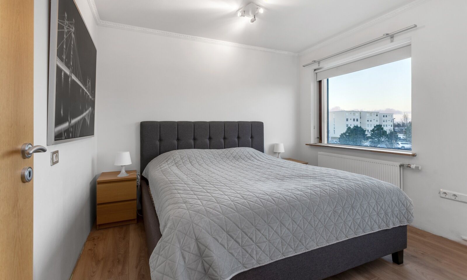 A bedroom with a bed, a nightstand, a window with a view, and a door.