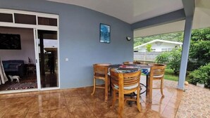 Ein überdachter Patio-Bereich mit Esstisch und Stühlen, eine Glastür zum Innenbereich und Blick auf ein Haus und viel Grün.