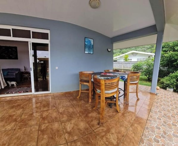A covered patio area with a dining table and chairs, a sliding glass door leading to an interior space, and a view of a house and greenery outside.