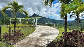 Ein geschwungener Betonweg führt zu einer weißen Pergola, umgeben von tropischen Pflanzen und Palmen, mit einem Bergpanorama im Hintergrund.