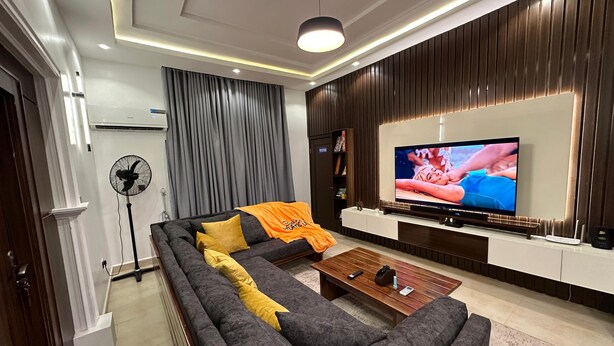 Una sala de estar moderna con un sofá modular, un televisor de pantalla plana, un ventilador de pie y una pared de acento de madera.