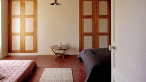 Une chambre à coucher avec un lit, une petite table ronde sur laquelle est posé un vase rose, un luminaire suspendu et une porte dont les panneaux sont en verre dépoli.