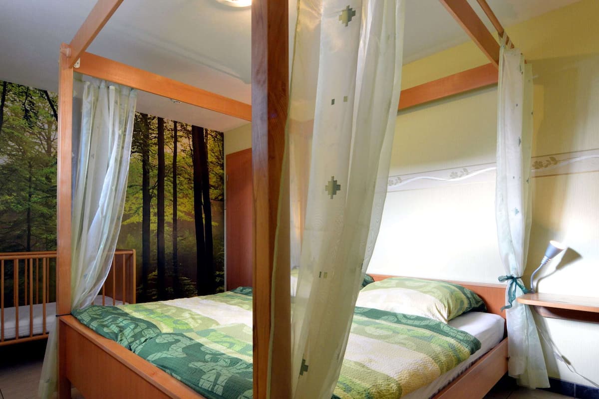 A four-poster bed with a forest mural backdrop, a wooden headboard, and a window with curtains.
