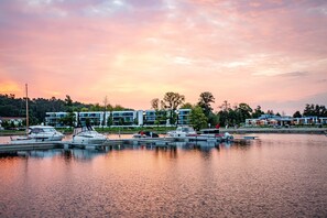 Un port de plaisance où sont amarrés plusieurs bateaux, un ensemble d’immeubles modernes en arrière-plan et une étendue d’eau calme qui reflète le ciel.