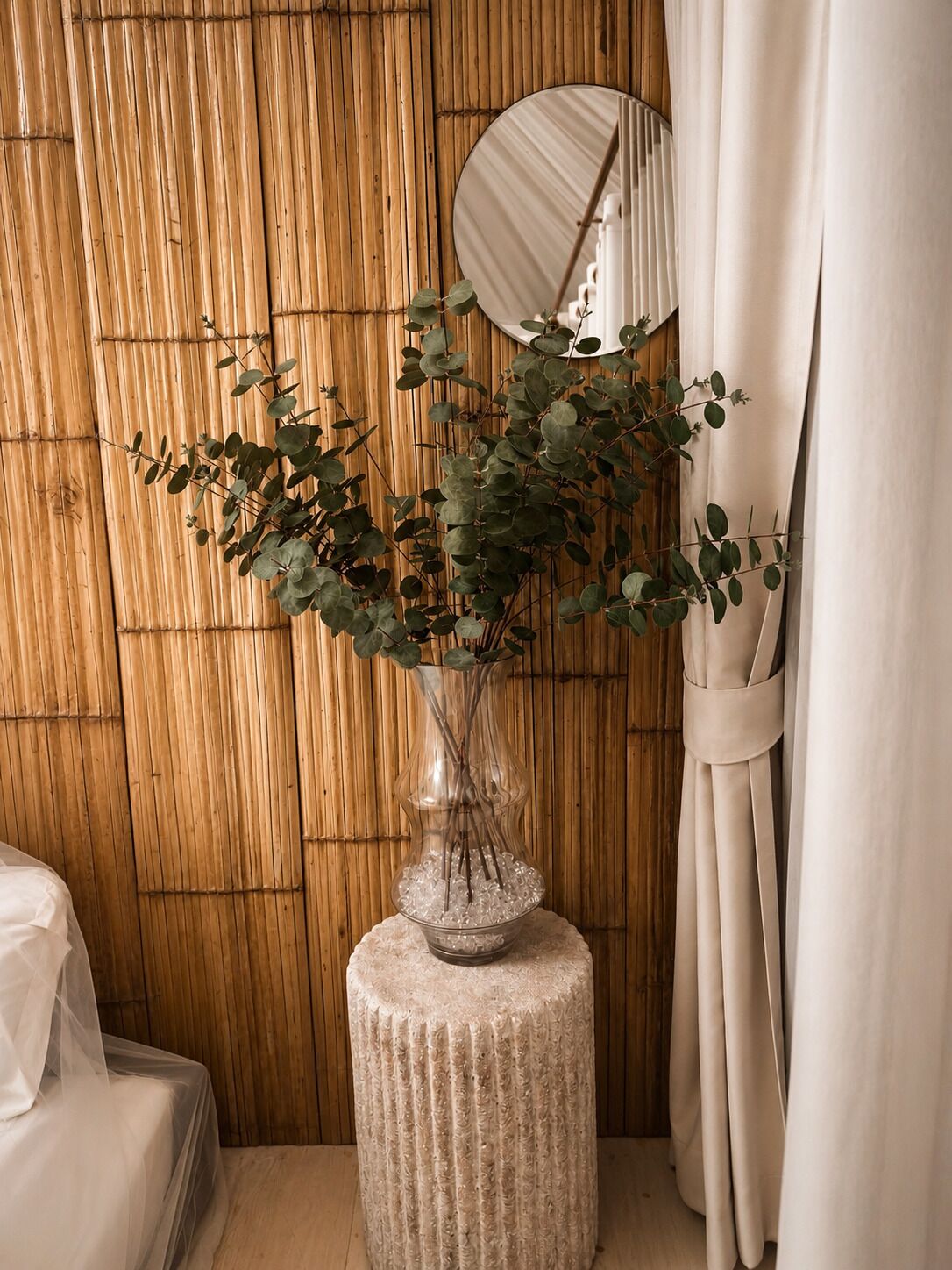A room with a wooden wall, a round mirror, a vase with green foliage, and a bed with a white bedspread.