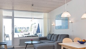 A modern living room with a grey sofa, a round table, and a dining table with chairs. There are pendant lights, a large window, and a view of the outside.