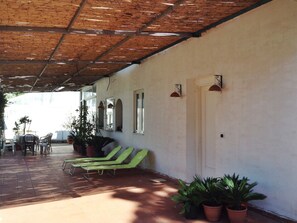 A covered patio area with a tiled floor, green lounge chairs, potted plants, and a white wall with windows and wall-mounted lights. - Apartment at the Foot of Mt. Vesuvius (Trecase)