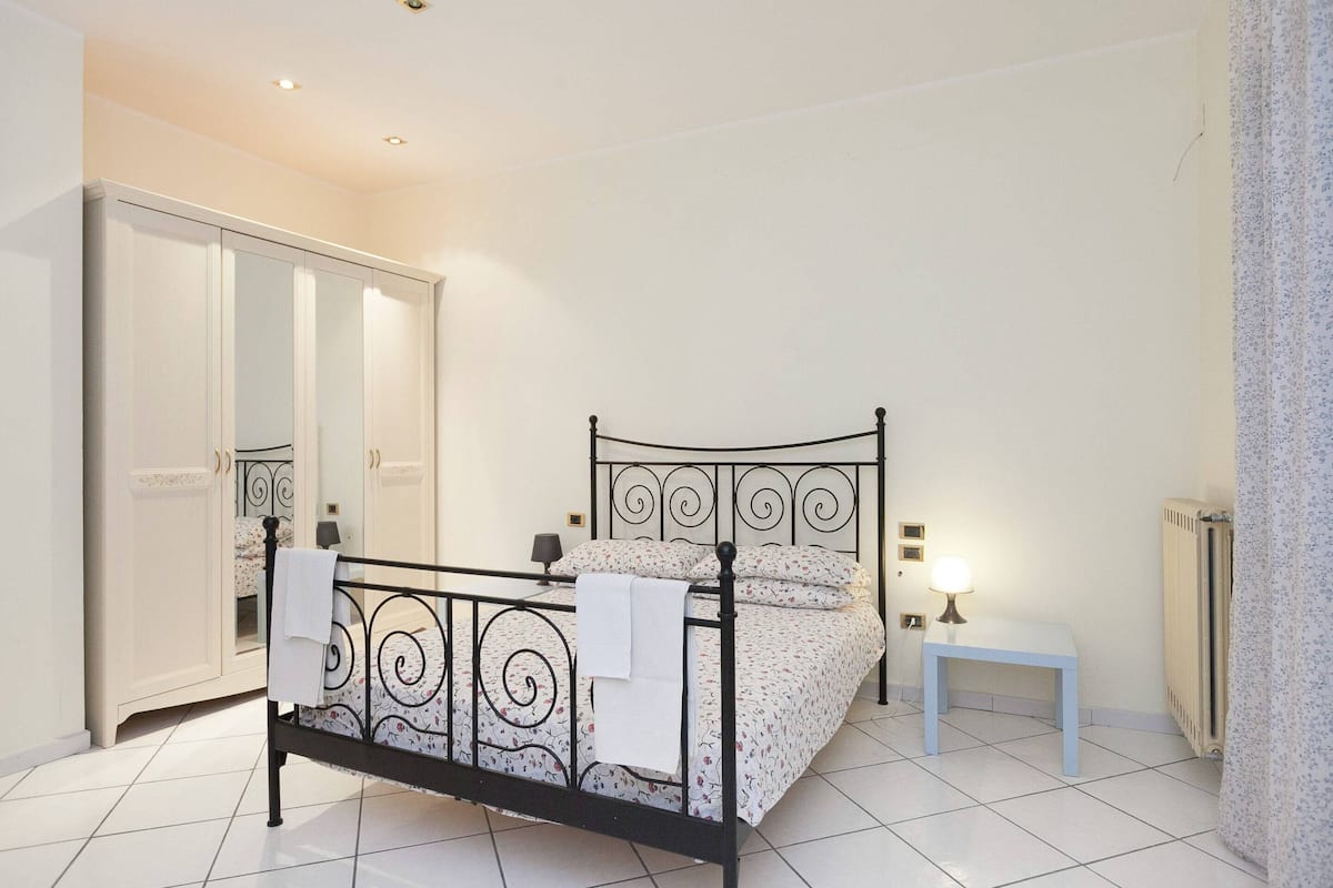 Un dormitorio con estructura de cama metálica, mesita de noche con lámpara, armario y radiador.