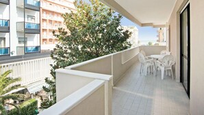 En balkon med hvide stole og et bord, med udsigt over bybillede med bygninger og et træ.