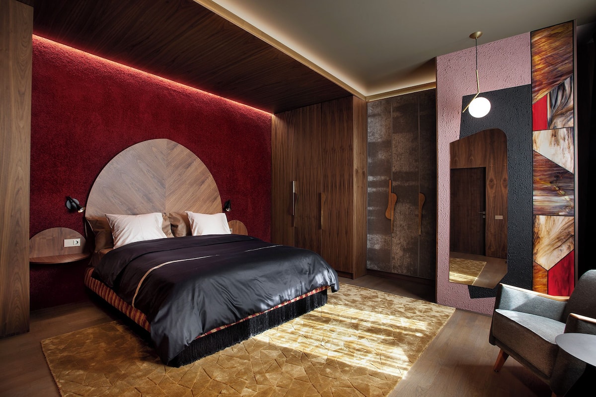 A modern bedroom with a large bed, a wooden headboard, a red accent wall, and a unique geometric artwork on the wall.