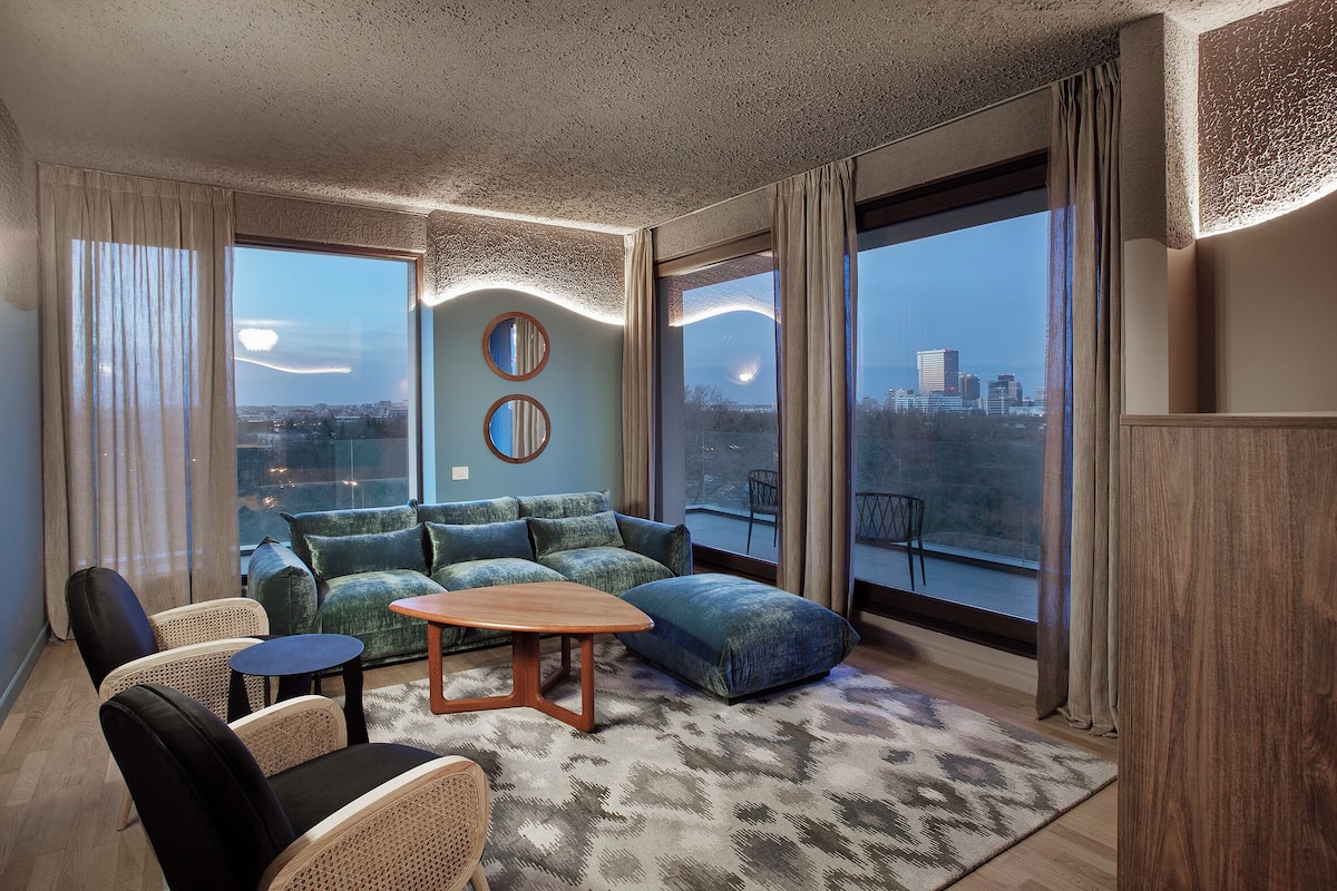 A modern living room with a sectional sofa, a round coffee table, and a view of the cityscape through large windows.