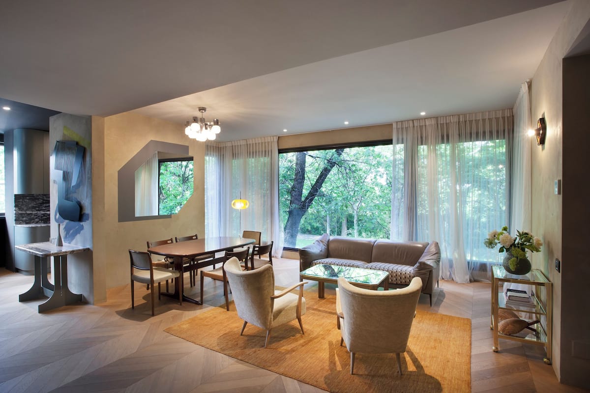 A modern living room with a dining area, large windows, and a view of trees.