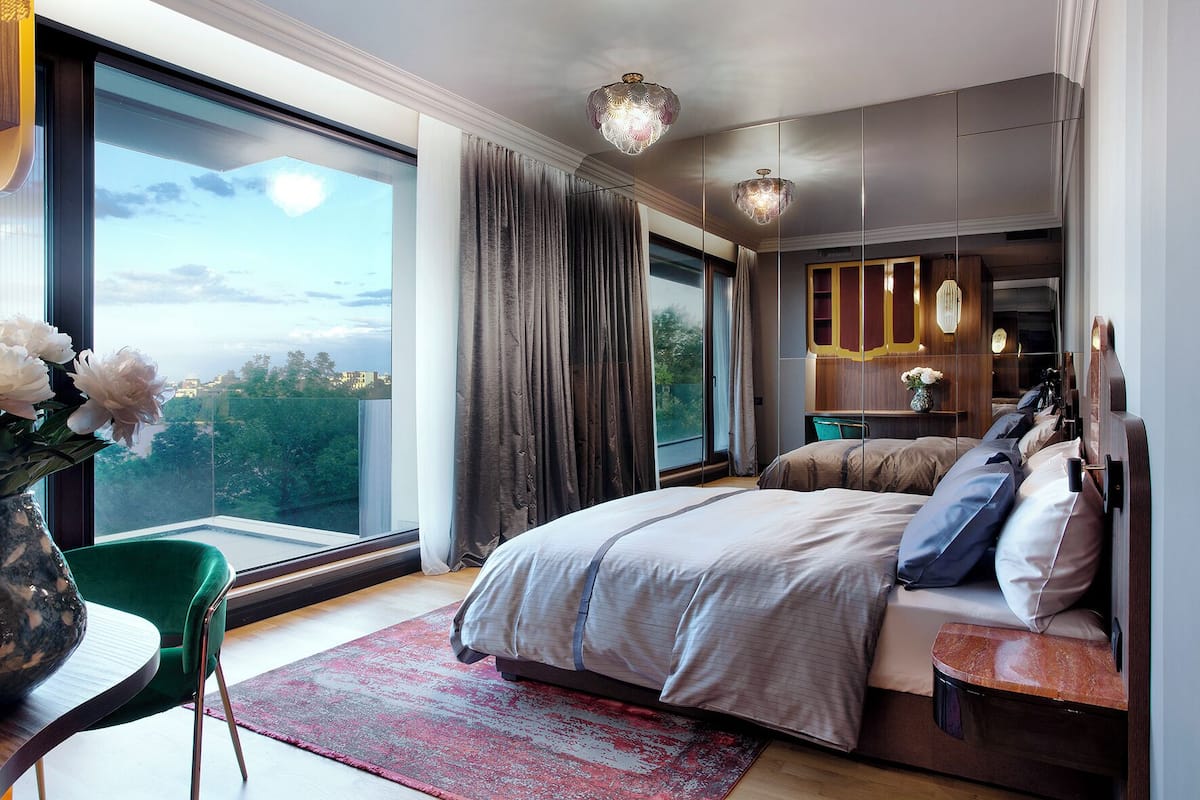 A modern bedroom with a large bed, a green chair, a desk, and a view of the outdoors.