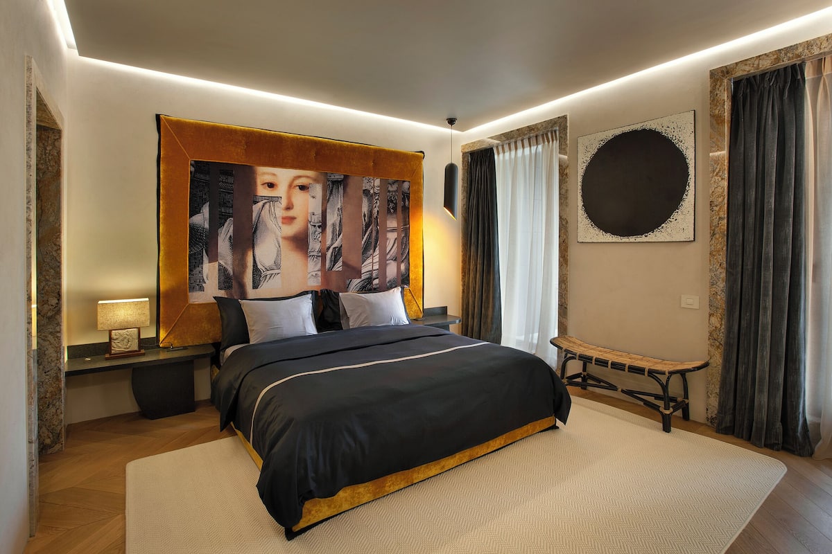 A modern bedroom with a large bed, a bench, a wall-mounted artwork, and a round mirror.