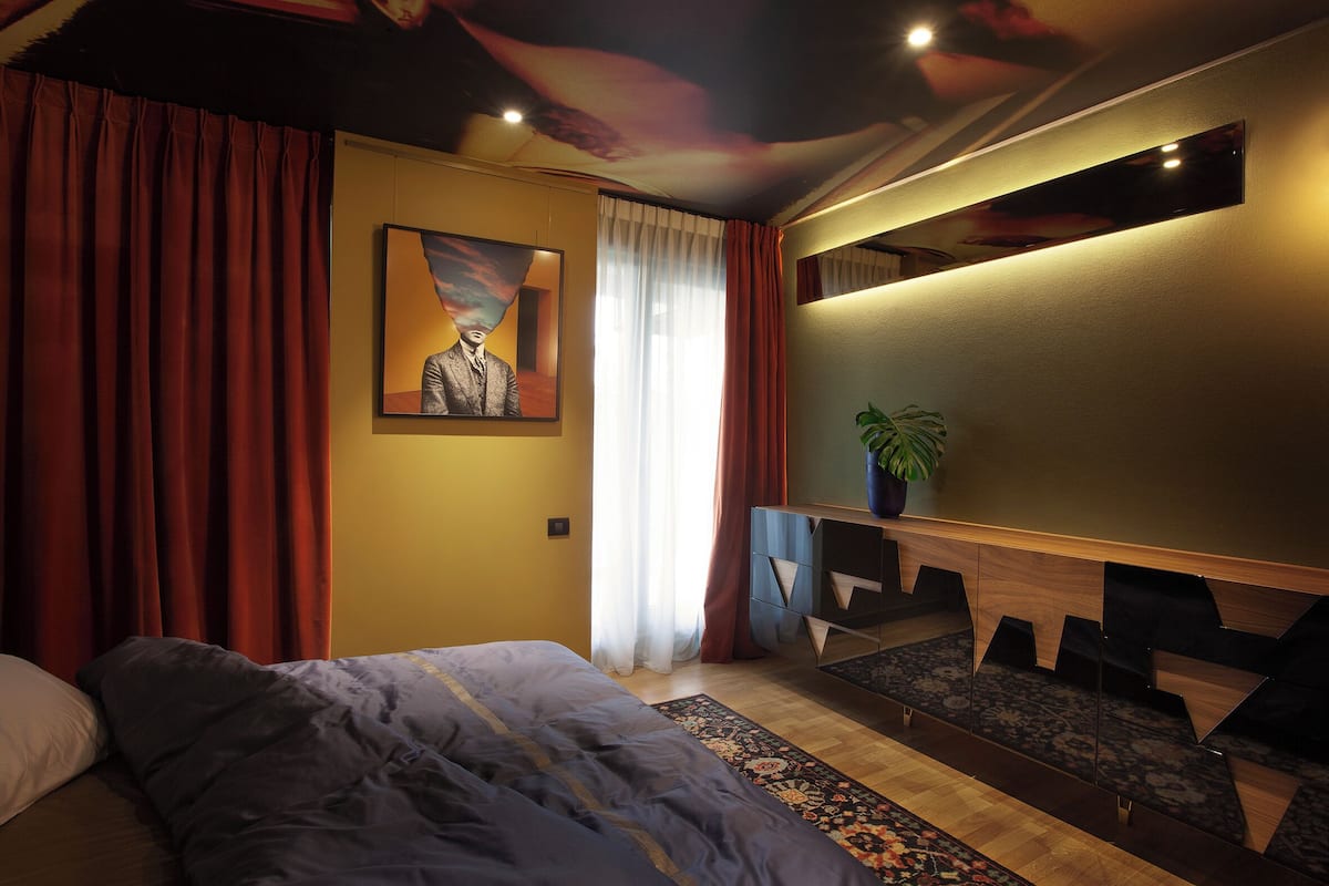 A bedroom with a bed, a desk, a chair, a plant, and a painting on the wall.