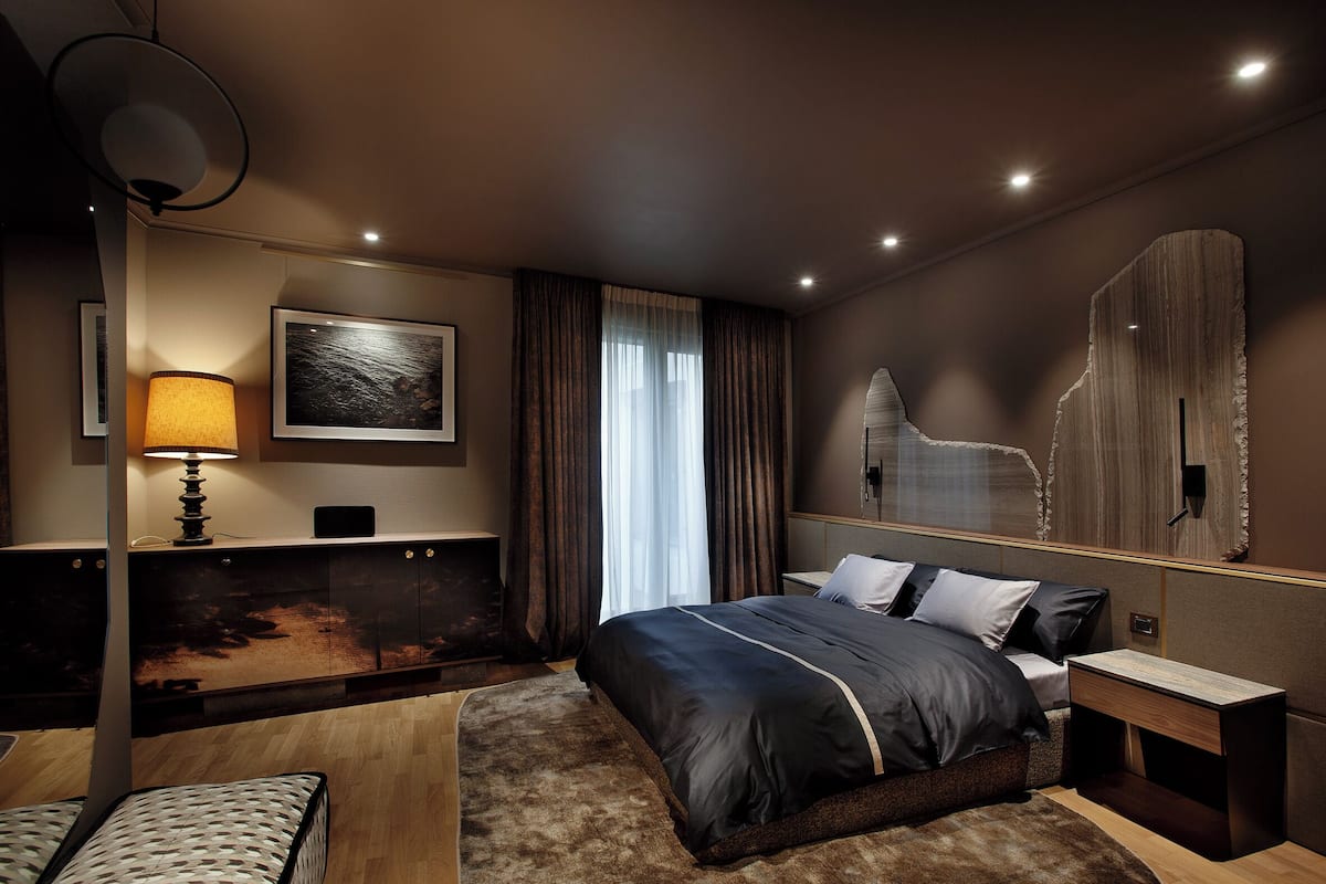 A modern hotel room with a large bed, a desk, a lamp, and a painting on the wall.