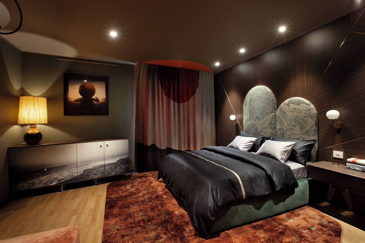 A modern bedroom with a large bed, a headboard with a unique design, a bedside table with a lamp, and a wall-mounted artwork.