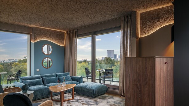 A modern living room with a blue sofa, wooden coffee table, and wicker chairs. It features large windows with a city view and a round mirror on the wall. - Room12 (Bucharest)