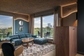 Una sala moderna con un sofá azul, una mesa de centro de madera y sillas de mimbre. Cuenta con ventanas amplias con vista a la ciudad y un espejo redondo en la pared.