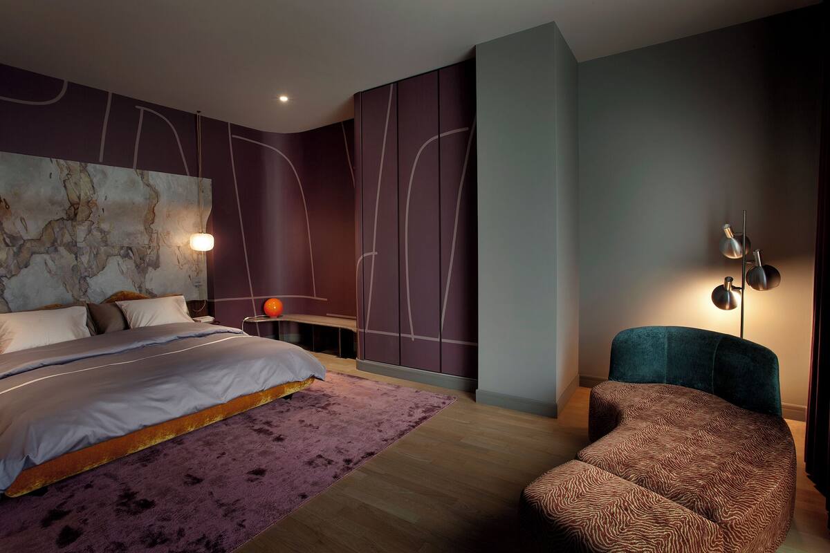 A modern bedroom with a bed, a side table, a chair, and a wall-mounted lamp.