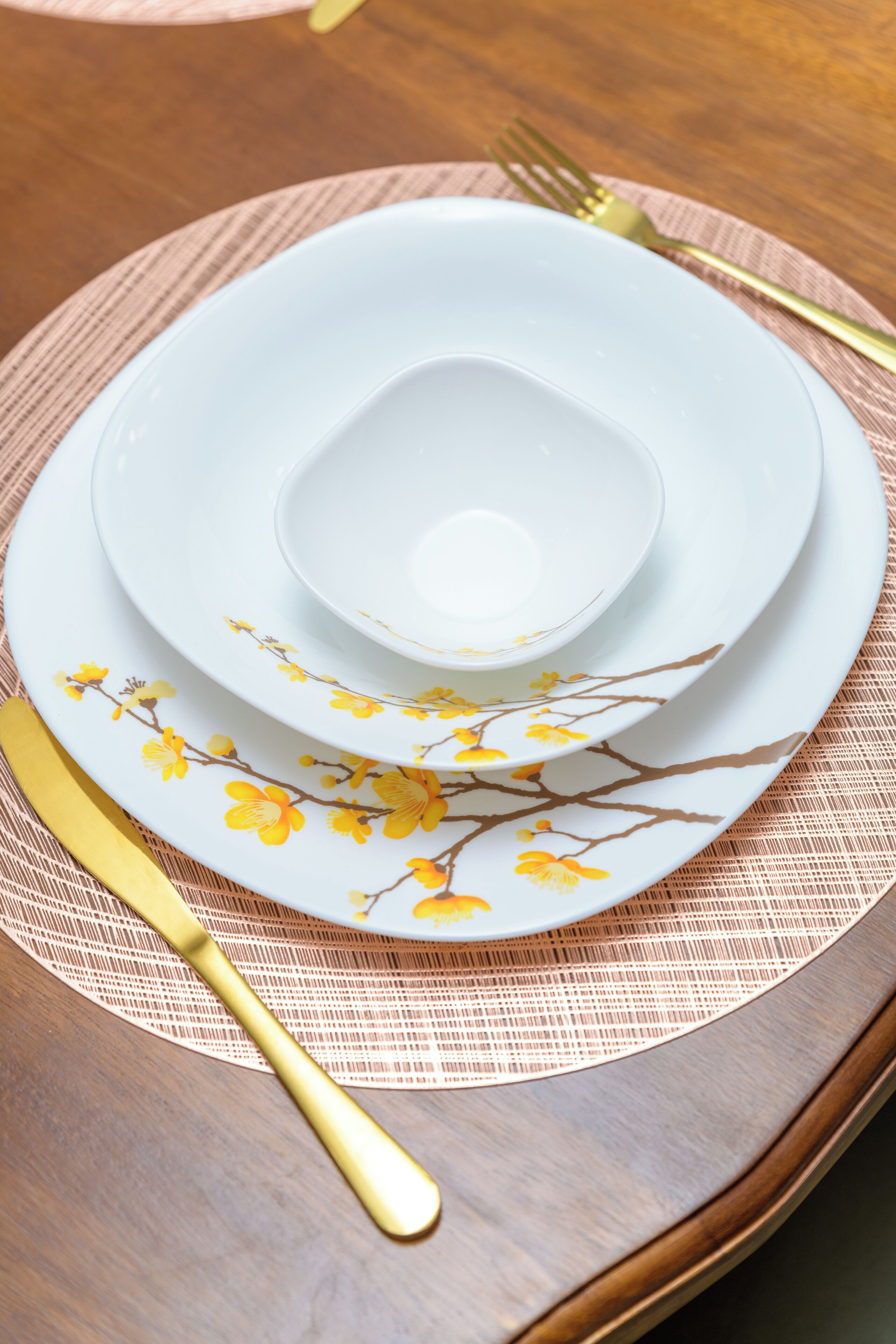 Una mesa dispuesta con platos blancos decorados con motivos florales amarillos, un tenedor dorado y un cuchillo dorado.