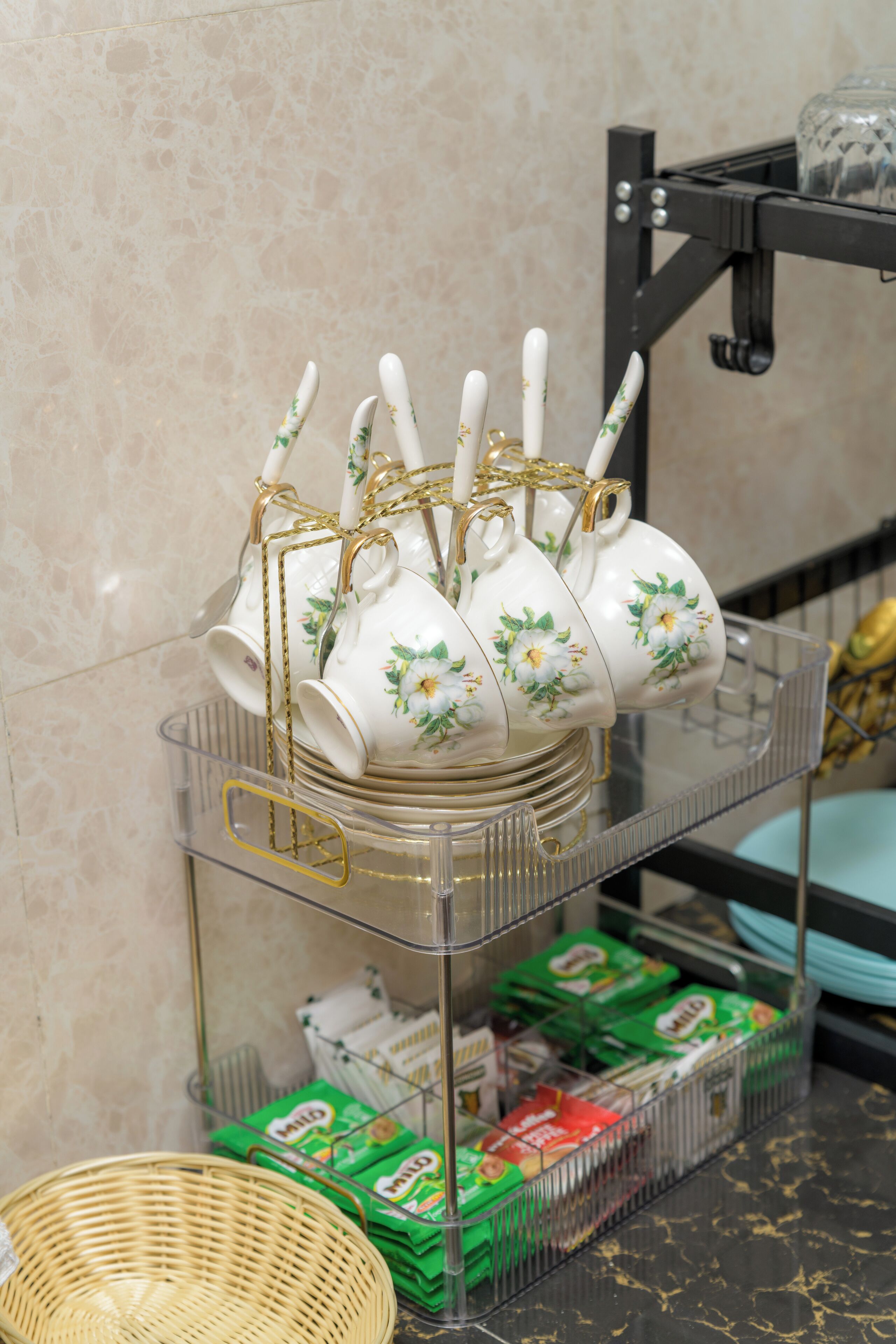 A kitchen area with a glass shelf holding white teapots and cups with floral designs, a wicker basket, and green cereal boxes.