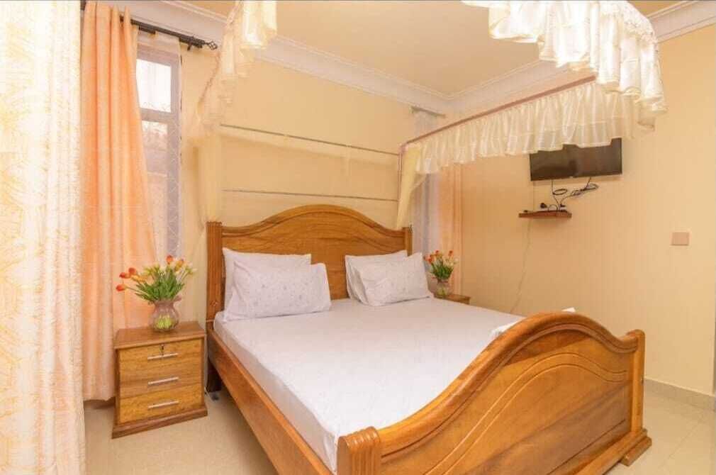 A bedroom with a wooden bed, a nightstand with drawers, a TV mounted on the wall, and a window with curtains.