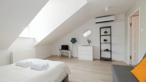 A modern bedroom with a sloped ceiling, a bed with white linens, a grey sofa, a desk with a computer, a mirror, and a small plant shelf. - ShortStayPoland Krakowskie Przedmieście (Lublin)