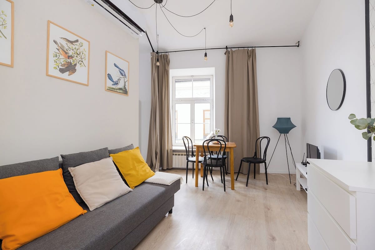 A modern living room with a grey sofa, yellow and white cushions, a dining table with chairs, a floor lamp, and a large window with curtains.