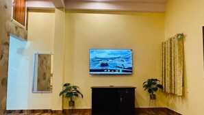 A room with a wooden floor, a flat-screen TV mounted on the wall, a black entertainment unit, and two potted plants. - Bhanu Bhawan (Jaipur)