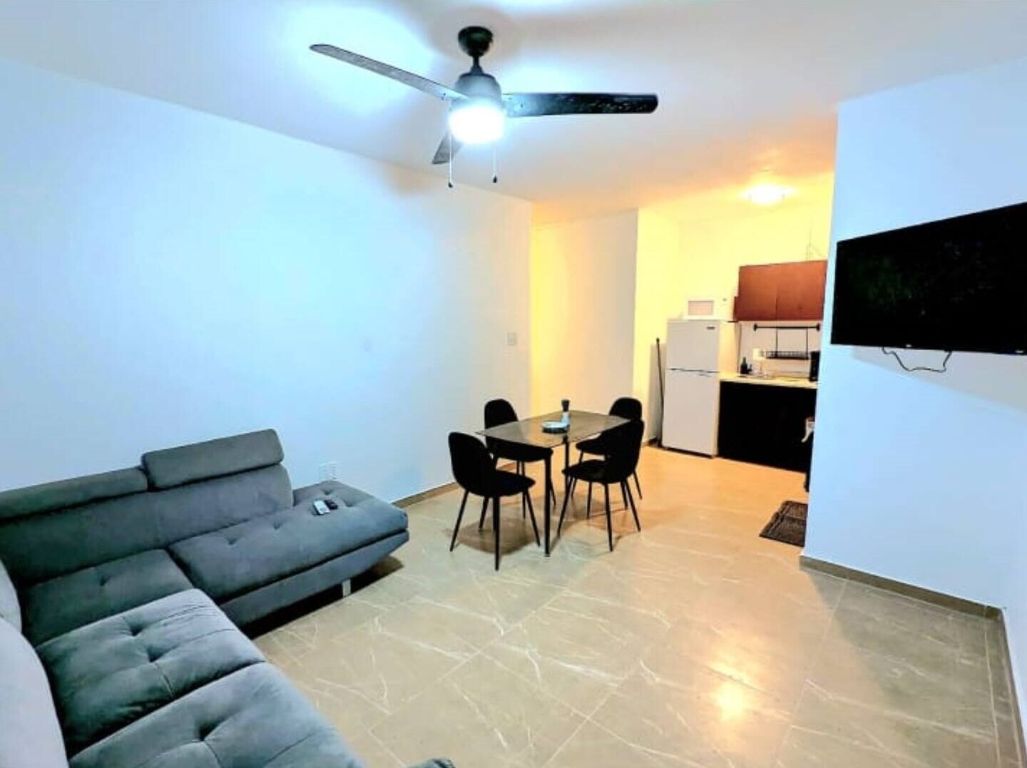 A compact living space with a sofa, a dining table with chairs, a ceiling fan, and a kitchen area with a refrigerator and stove.