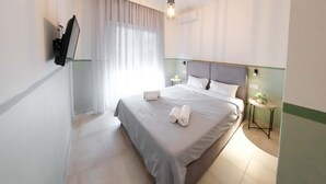 A neatly made bed with a grey bedspread and white pillows, a bedside table with a lamp, and a window with sheer curtains. - Orsiida Classic Double Room Volos (Volos)