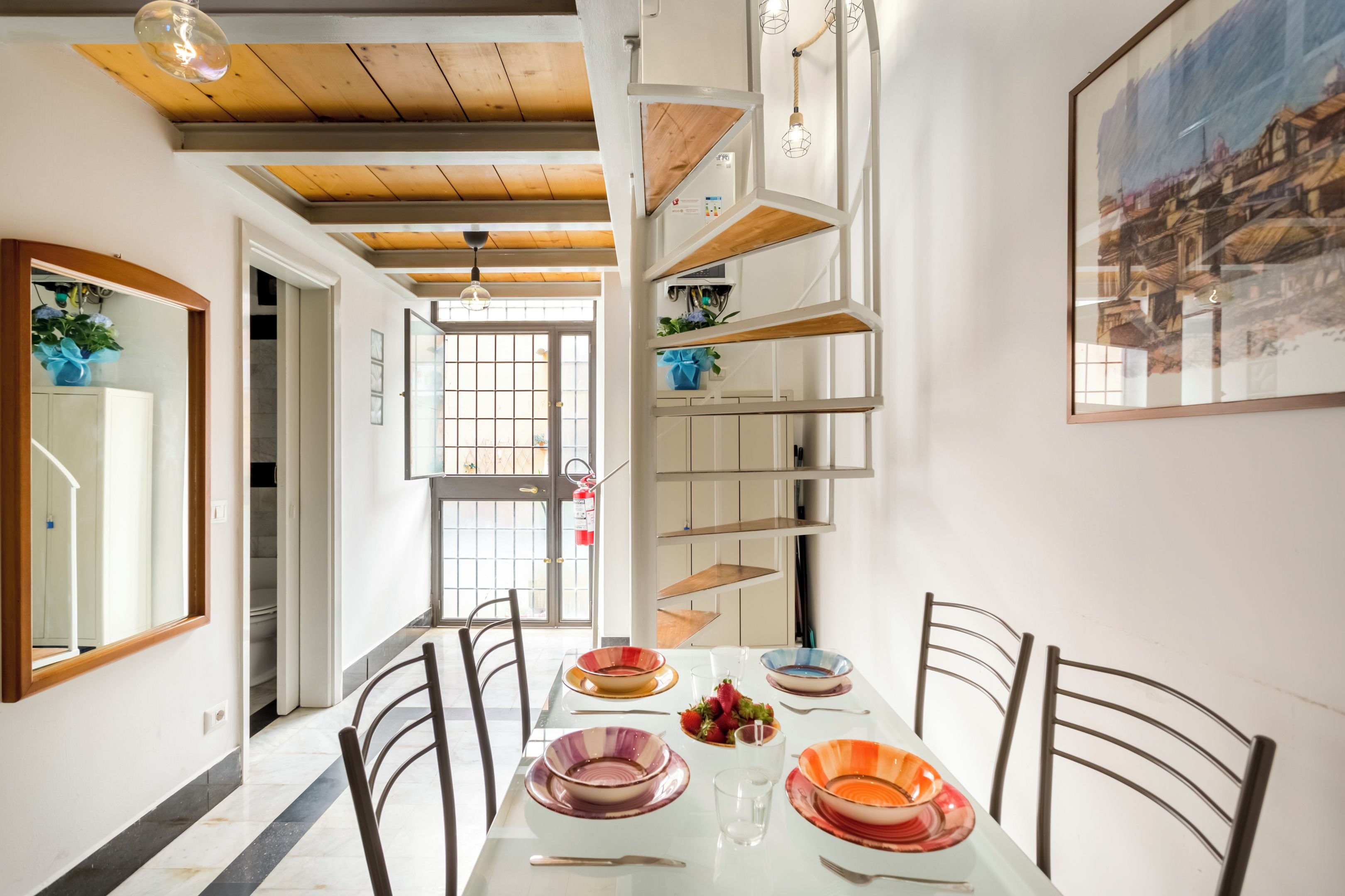 Zona pranzo con tavolo bianco, piatti colorati e sedie. Si vede una scala con scaffali e un quadro appeso al muro.