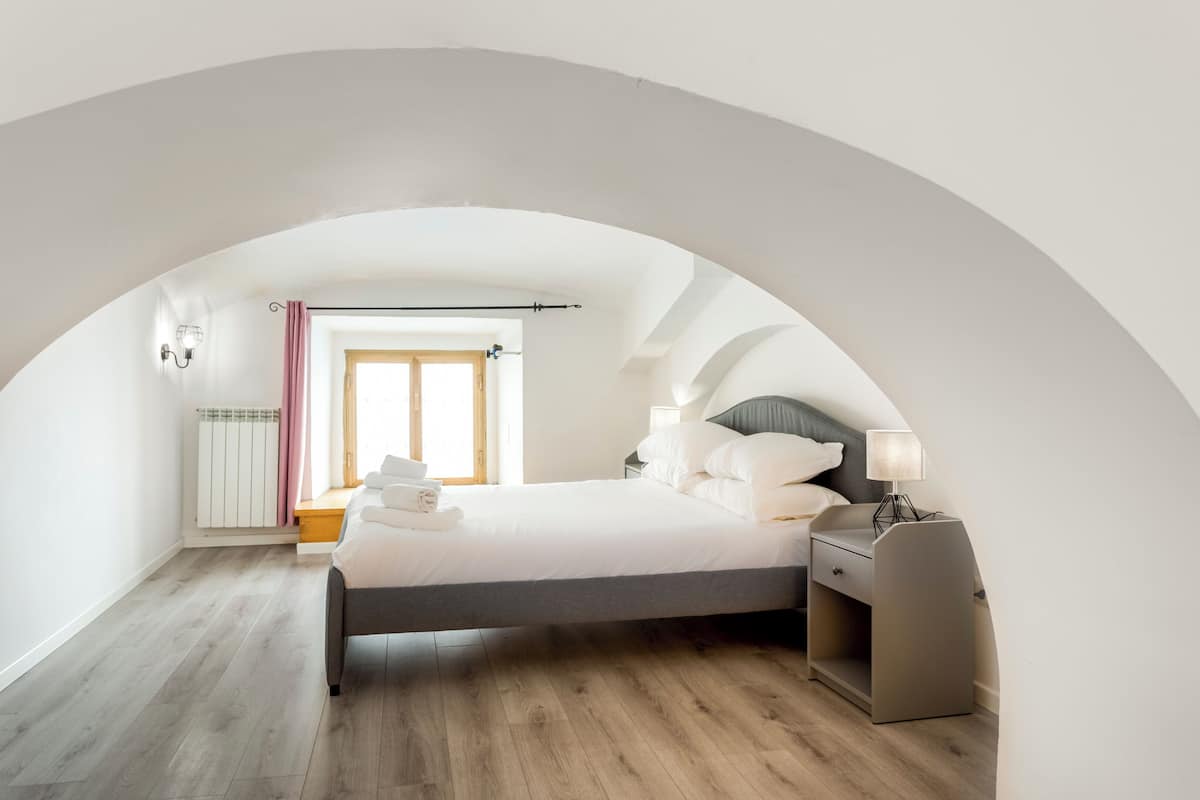 A bedroom with a bed, bedside table, lamp, and a view through an arched doorway.