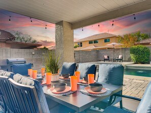 An outdoor patio with a dining table set for four, surrounded by chairs with cushions. There is a grill and a pool in the background. - Dream Home in Gilbert! Fun Times, Lots of Amenities! Pool Home. (Gilbert)