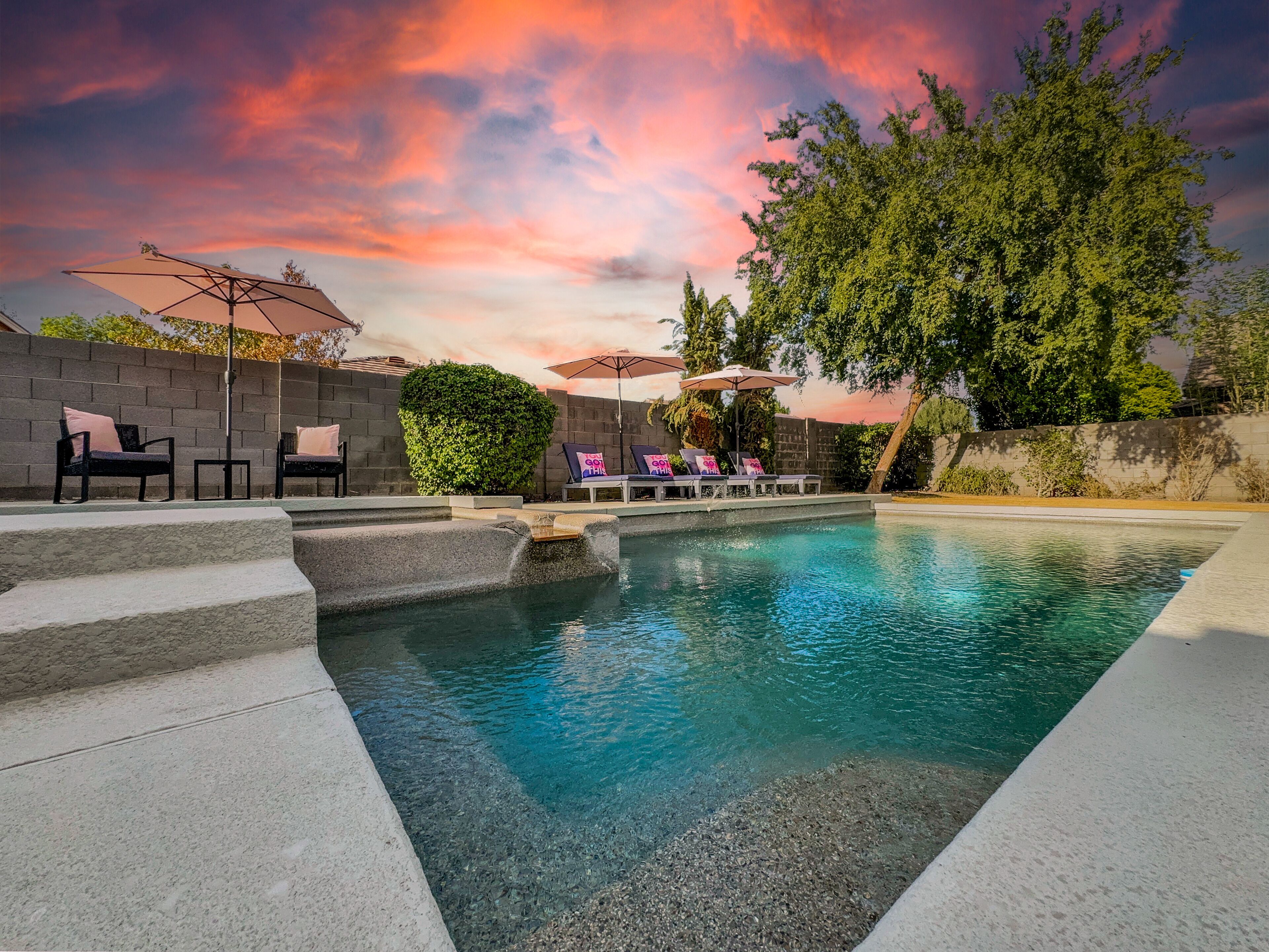 A swimming pool with lounge chairs and umbrellas, surrounded by a stone wall and trees.