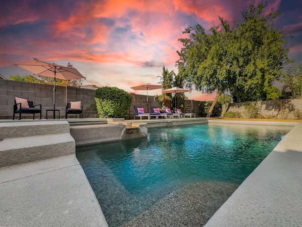 A swimming pool with lounge chairs and umbrellas, surrounded by a stone wall and trees. - Dream Home in Gilbert! Fun Times, Lots of Amenities! Pool Home. (Gilbert)