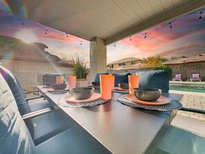 An outdoor dining area with a pool, string lights, and a sunset view.