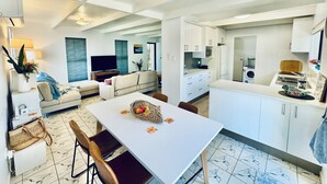 A modern kitchen with white cabinets, a dining table with chairs, a TV, and a sofa in the living area.