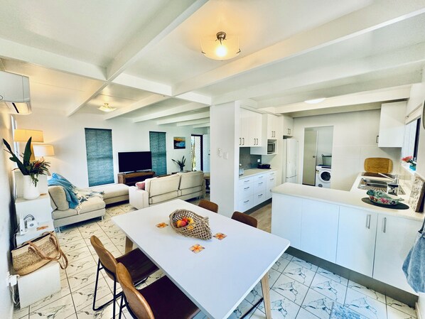 Una cocina moderna con armarios blancos, una mesa de comedor con sillas, un televisor y un sofá en la sala.