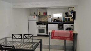 A compact kitchen with a refrigerator, stove, microwave, and washing machine. There is a glass table with chairs and a red tablecloth. - 1 bedroom house 800m from the sea (Saint-Raphaël)