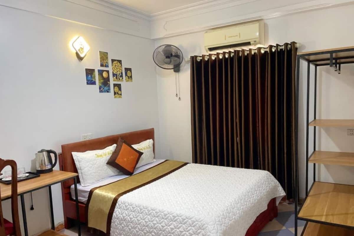 Une chambre d’hôtel bien rangée, avec un lit, un bureau, une chaise, un radiateur et un ventilateur fixé au mur.