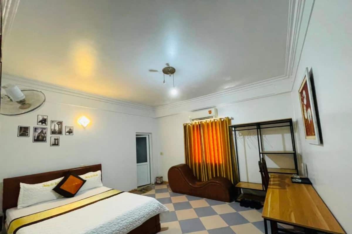 Une chambre d’hôtel équipée d’un lit, d’un bureau, d’une chaise, d’un ventilateur de plafond et d’une fenêtre avec des rideaux.