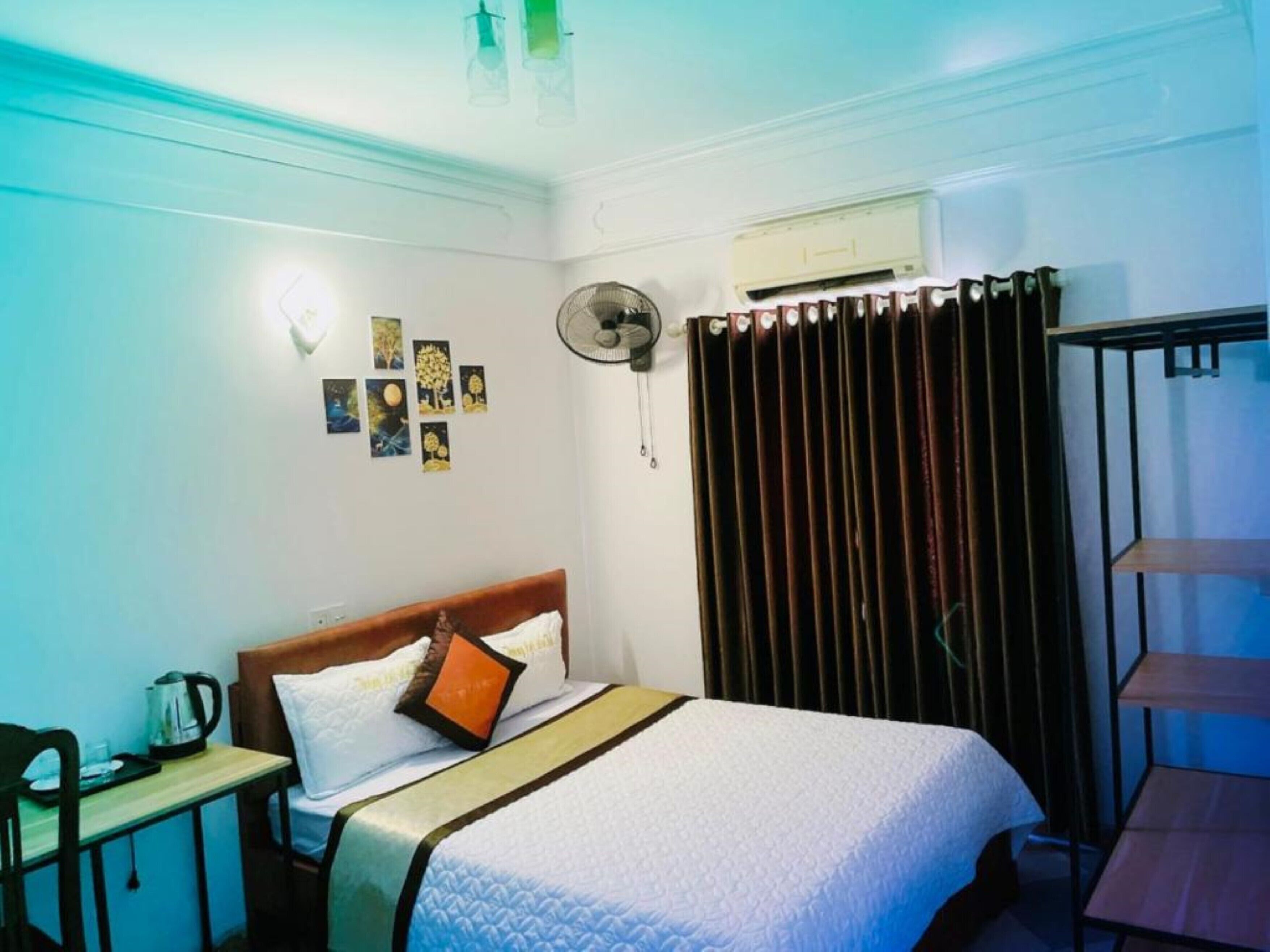 Quarto de hotel com cama, mesa com chaleira, ventilador e cortina.