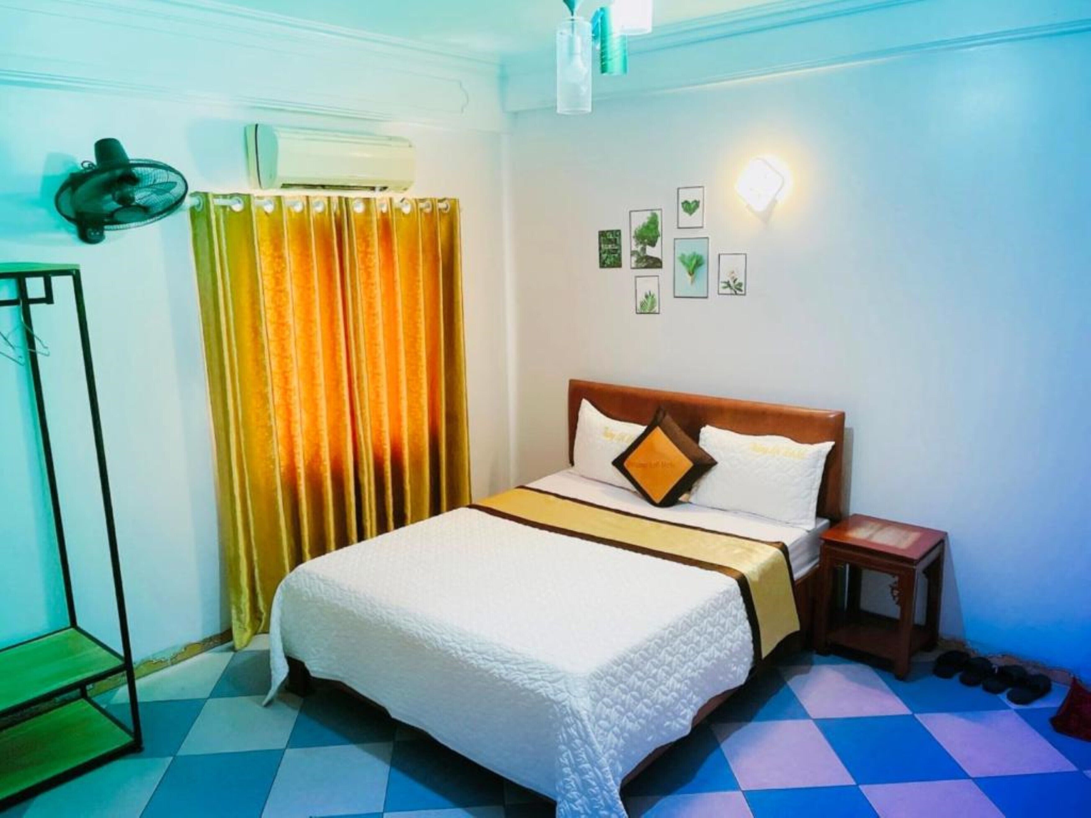 A hotel room with a bed, a nightstand, a mirror, a fan, a window with curtains, and wall art.