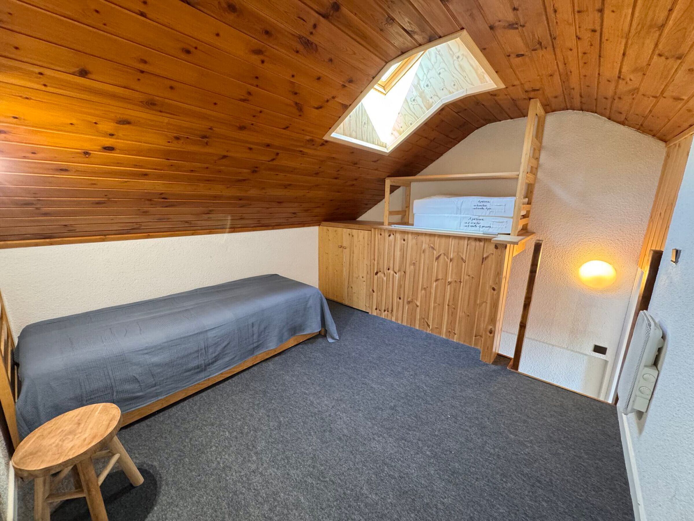 Ett litet, ombonat rum med en våningssäng, en enkelsäng, en träpall, ett takfönster och en vit vägg med en lampa.