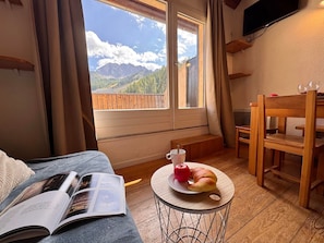 A room with a bed, a table with a croissant and apple, a book, and a view of mountains through a window. - Apartment at the Slopes near ESF (Vars)